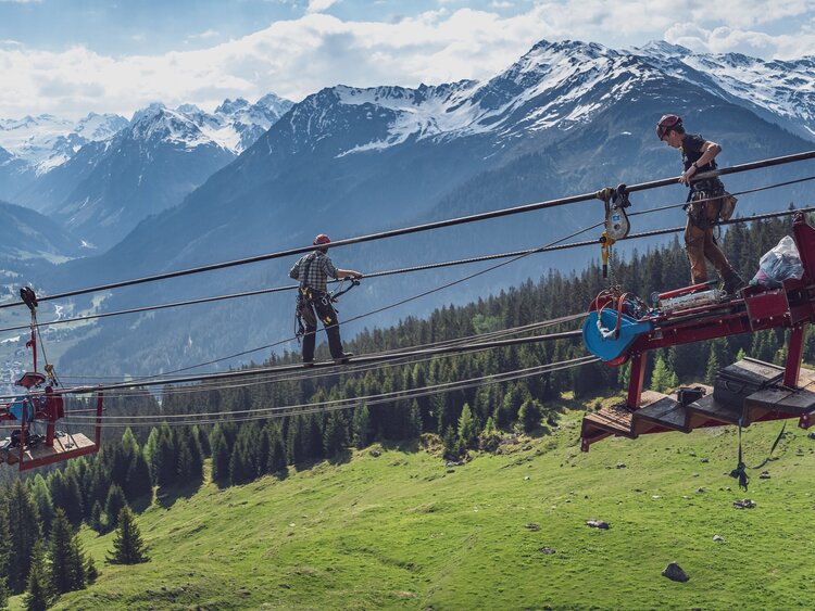 Zwei Arbeiter klettern auf dem Tragseil zur sogenannten Melkmaschine. | © Davos Klosters Mountains