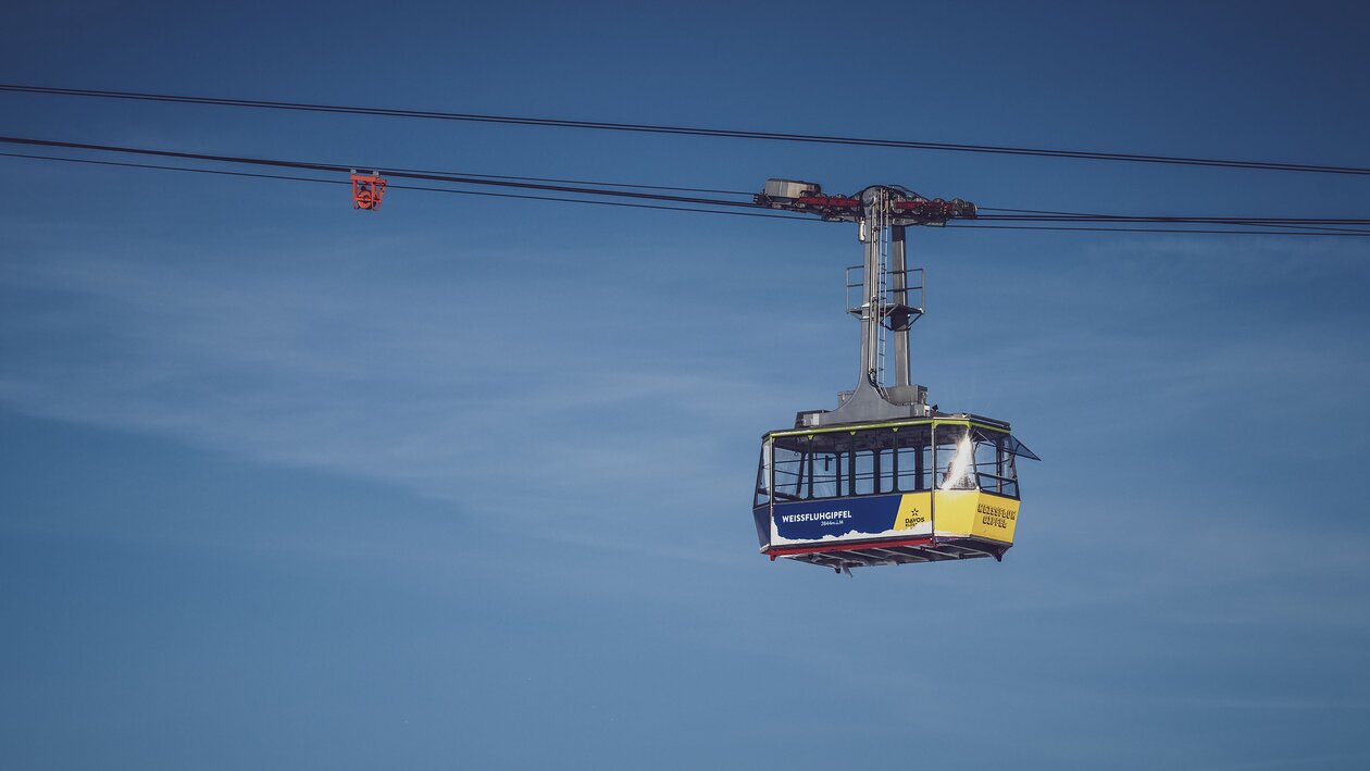 Vor blauem Himmel schwebt die blau/gelbe Gondel der Gipfelbahn zum Weissfluhgipfel auf Parsenn.   | © Davos Klosters Mountains