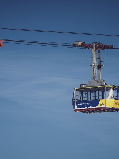 Vor blauem Himmel schwebt die blau/gelbe Gondel der Gipfelbahn zum Weissfluhgipfel auf Parsenn.   | © Davos Klosters Mountains