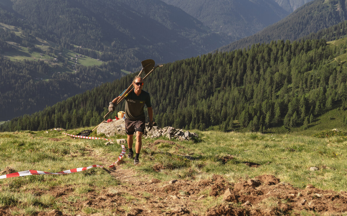 Christoph Koch, Mitglied der Trail Crew Davos, ist mit Rechen und Schaufel über der Schulter auf dem Berg unterwegs.  | © Carmen Wenger / Destination Davos Klosters