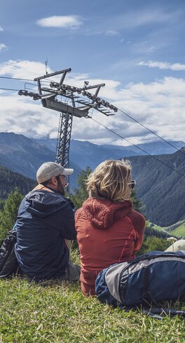 Eine Familie sitzt auf der Bergwiese auf dem Rinerhorn und geniesst die Natur, dahinter schwebt eine Gondel der Rinerhornbahn in Richtung Berg.  | © Davos Klosters Mountains