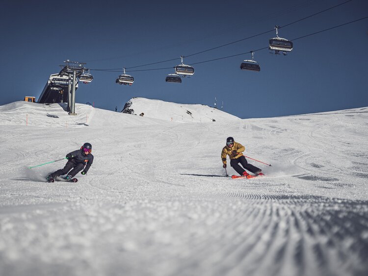 Eine Skifahrerin und ein Skifahrer fahren über die Skipiste auf Parsenn, der Sessellift Rapid im Hintergrund. | © Davos Klosters Mountains